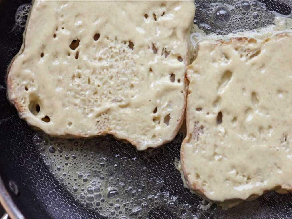 Two slices of bread coated in a batter are being cooked in a frying pan with melted butter, likely in the process of making French toast.